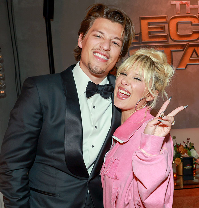 Young woman in pink jacket smiling and posing with man in black tuxedo at an event, related to Millie Bobby Brown adoption news. Young woman in pink jacket smiling and posing with man in black tuxedo at an event, related to Millie Bobby Brown adoption news.
