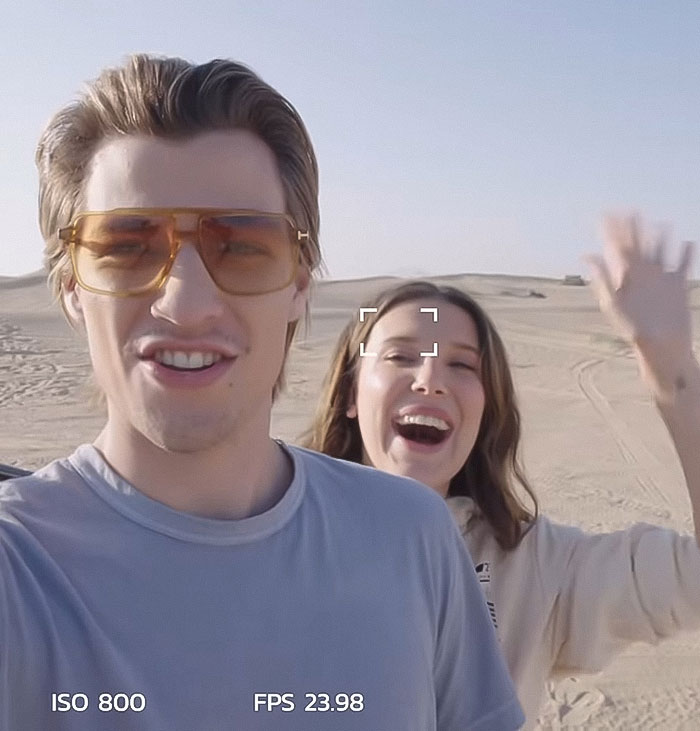 Young man wearing brown tinted glasses and woman waving happily outdoors, related to Millie Bobby Brown adoption news. Young man wearing brown tinted glasses and woman waving happily outdoors, related to Millie Bobby Brown adoption news.