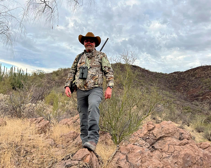 Male trophy hunter in camo gear and hat stands on rocky terrain during safari, highlighting millionaire hunter karma incident. Male trophy hunter in camo gear and hat stands on rocky terrain during safari, highlighting millionaire hunter karma incident.