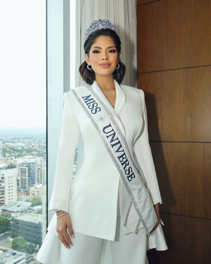 Miss Universe winner posing indoors wearing crown and white suit with sash, standing near window with city view background.