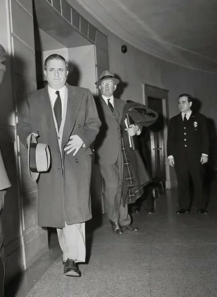Vintage black and white photo of a mob boss walking with companions and a police officer in a building hallway, notorious mob bosses.