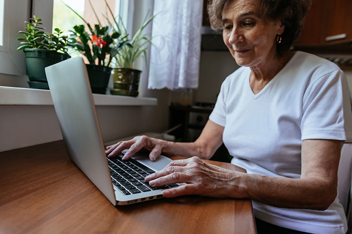 Older woman using laptop at home near plants, highlighting mom disabling MIL’s baby cam access for privacy concerns. Older woman using laptop at home near plants, highlighting mom disabling MIL’s baby cam access for privacy concerns.