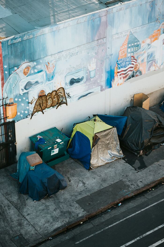 Street tents and graffiti mural highlighting performative behaviors in society near urban sidewalk.
