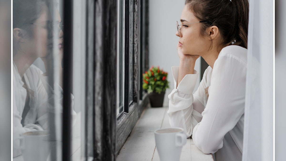 Woman sitting alone by window, looking sad and reflective, illustrating a cheating and shocking confrontation scenario.