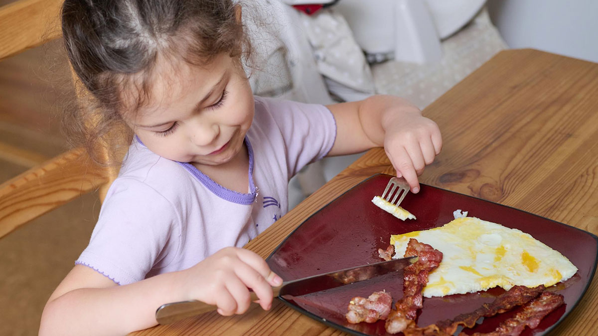 Young child eating meat and eggs at a wooden table despite being a vegan child fed by neighbour.