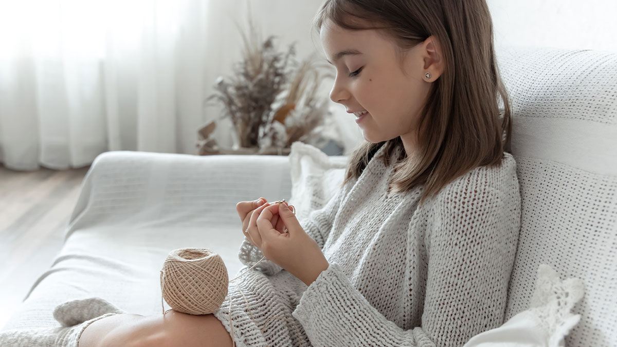 Young girl smiling while knitting with yarn on a white couch, showing a peaceful home moment with grieving family dynamics.