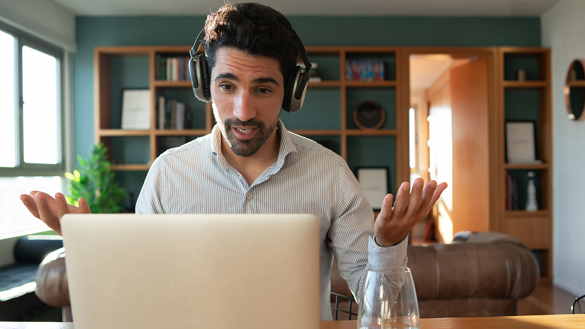 Man with headphones in home office video calling coworkers, refusing to change surname for American coworkers.