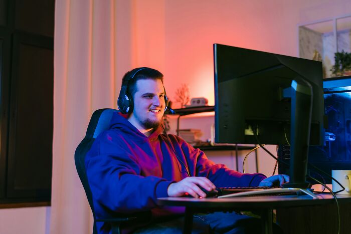 Young man in a hoodie and headset shopping online using a desktop computer in a cozy, warmly lit room.