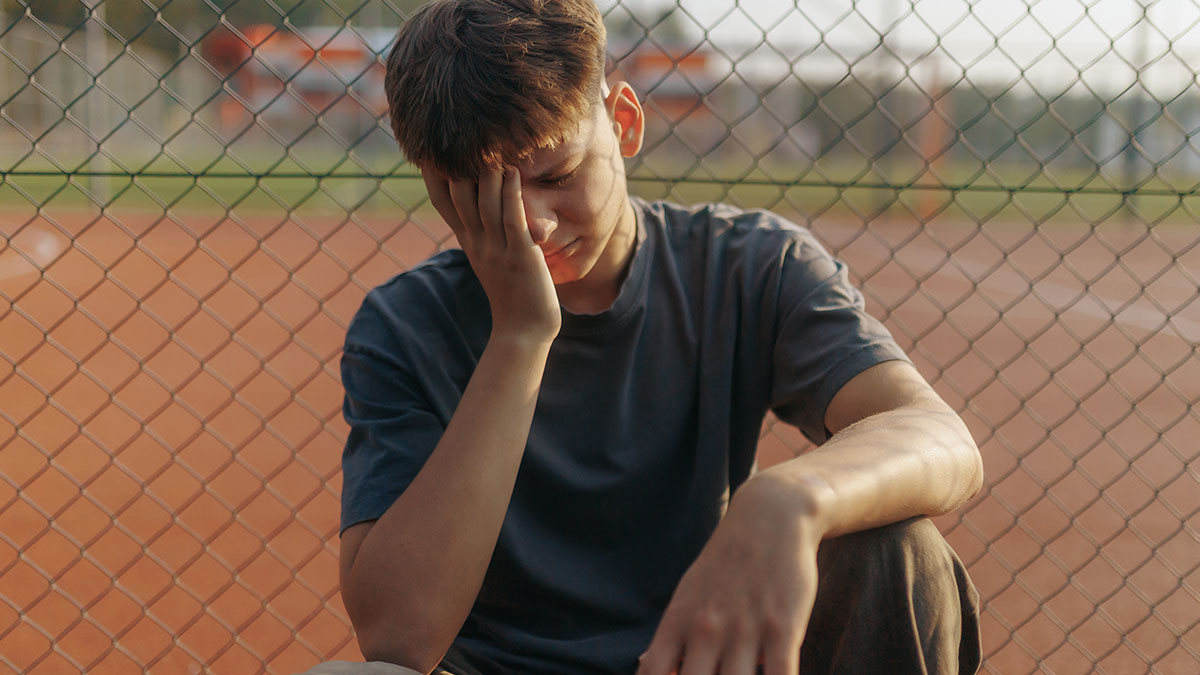 Teen boy sitting by chain-link fence, looking upset and neglected, reflecting parents forgetting the one kid they had.