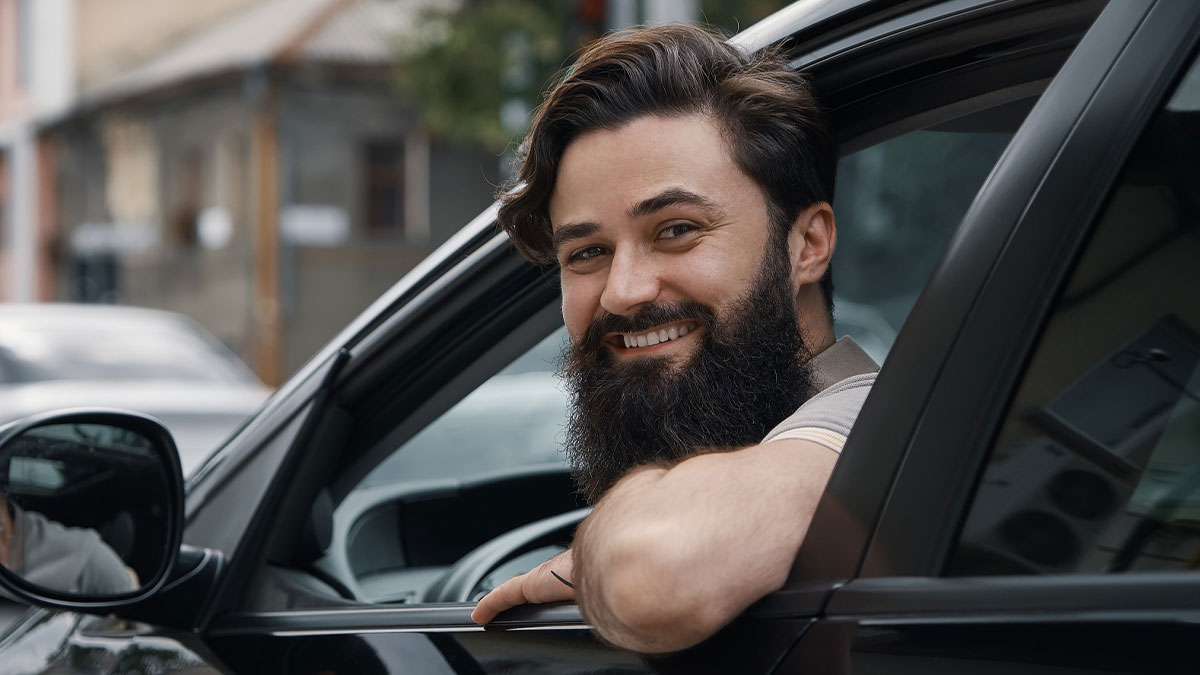 Bearded man smiling from car window after blocking another car's driver door by parking too close in a tight spot.