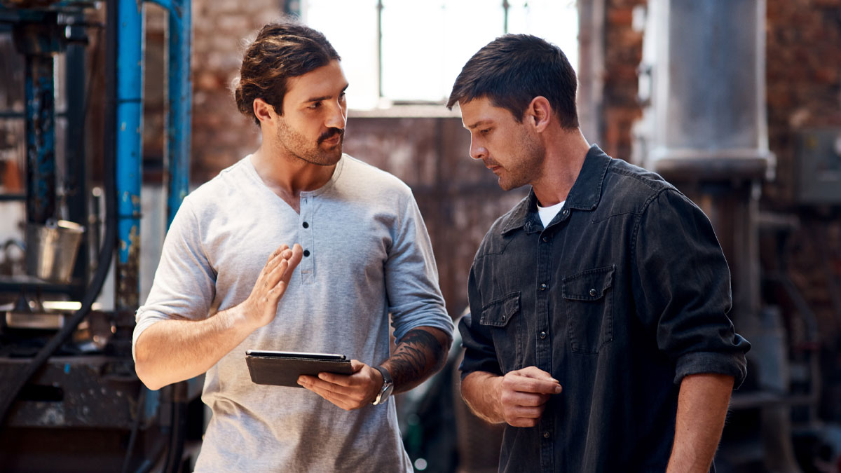 Two men discussing profession secrets using a tablet in an industrial workshop setting with focused expressions.