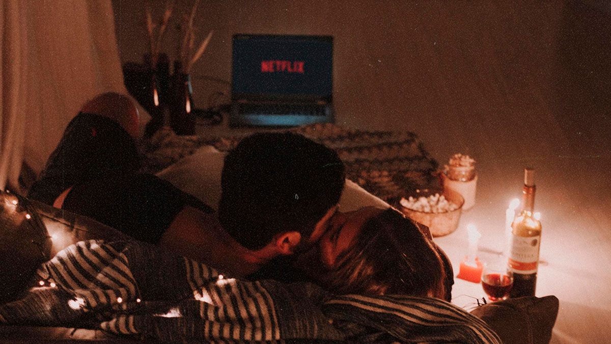 Couple lying on bed with fairy lights, sharing a kiss in a cozy room with Netflix on laptop and wine bottle nearby.
