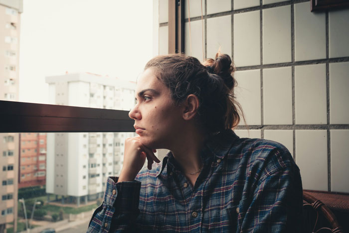 Woman in a plaid shirt looking out window with a pensive expression, reflecting on family vacation stress and emotions. Woman in a plaid shirt looking out window with a pensive expression, reflecting on family vacation stress and emotions.