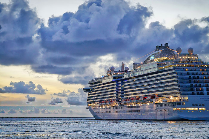Large cruise ship sailing on calm ocean waters under a cloudy sky at sunset with bright deck lights visible.