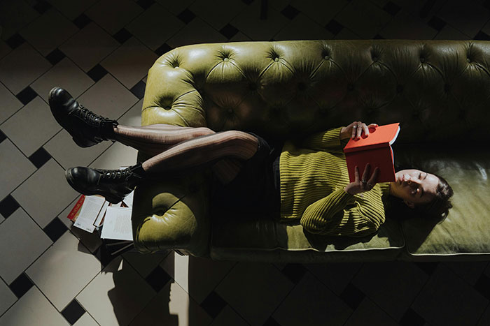 Young woman lying on green couch holding red book, wearing striped tights and black boots in dimly lit room. Young woman lying on green couch holding red book, wearing striped tights and black boots in dimly lit room.