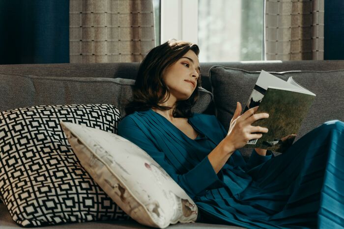 Woman in blue dress relaxing on couch by window, reading a book, illustrating picking the wrong line at the grocery store concept.