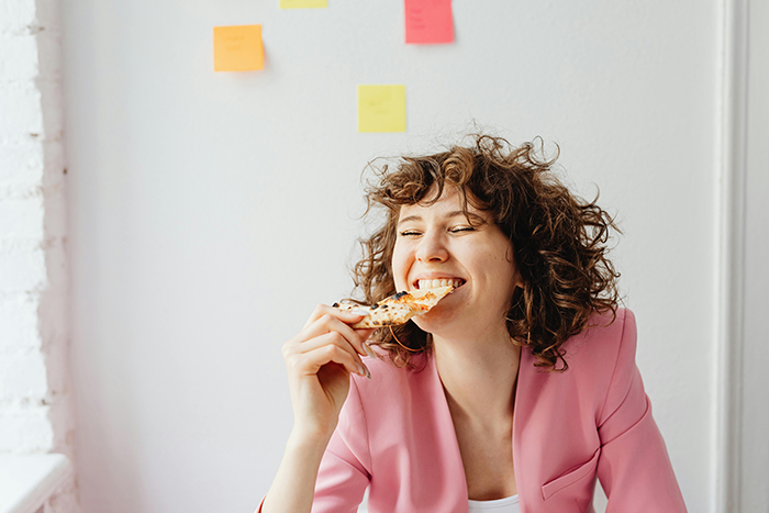 Woman eating pizza during lunch break, smiling happily in an office setting with colorful sticky notes on the wall.