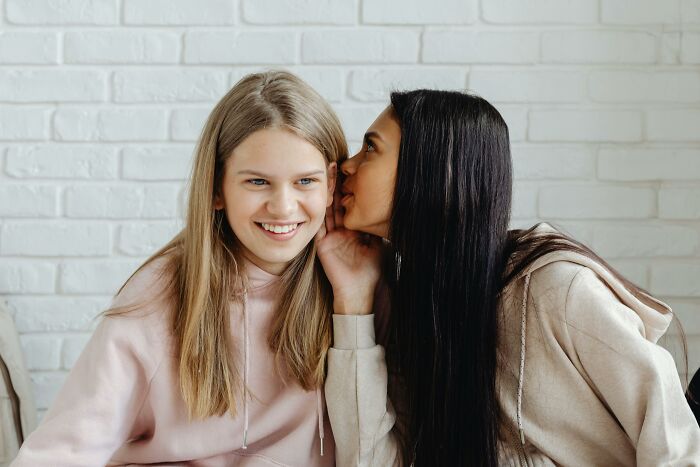 Two young women in casual hoodies share a secret, illustrating picking the wrong line at the grocery store experience.