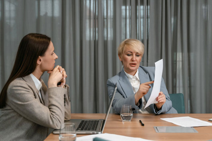 Boss in gray suit pointing at document while employee in beige blazer listens during tense office meeting Boss in gray suit pointing at document while employee in beige blazer listens during tense office meeting