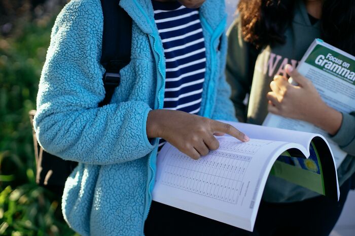 Two students outdoors studying grammar and sentence errors, one pointing to a page in an open workbook. Two students outdoors studying grammar and sentence errors, one pointing to a page in an open workbook.