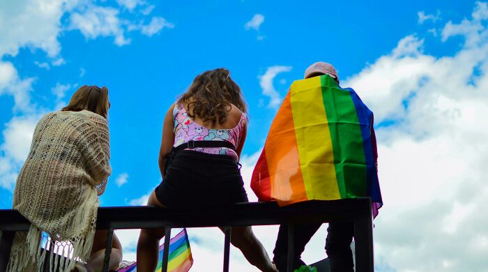 Three people sitting outdoors against a blue sky, one wearing a rainbow flag representing performative behaviors in society.