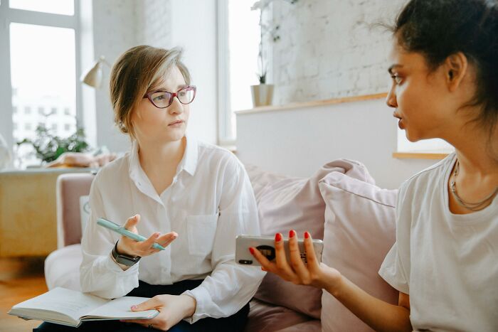 Two women discussing misheard phrases, one with glasses holding a notebook, in a bright modern living room. Two women discussing misheard phrases, one with glasses holding a notebook, in a bright modern living room.