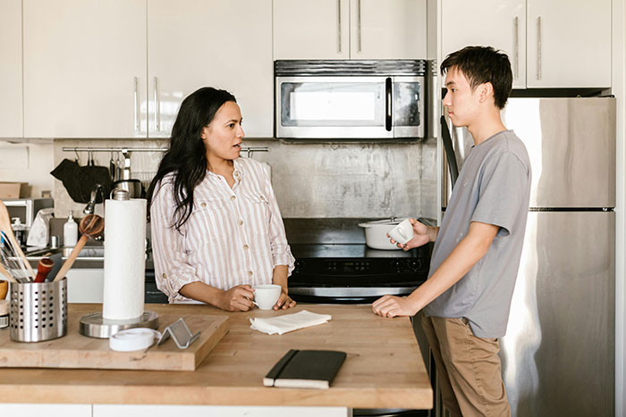 Woman and roommate in kitchen discussing cat duties as she plans family vacation, roommate refusing litter box responsibilities.