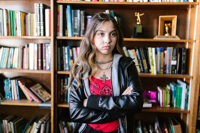 Teen girl with crossed arms looks upset in front of a bookshelf, reflecting challenges of winning over fiancee’s kids. Teen girl with crossed arms looks upset in front of a bookshelf, reflecting challenges of winning over fiancee’s kids.