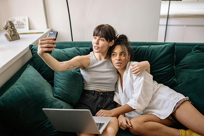 Two women sitting closely on a sofa taking a selfie, reflecting a moment before dinner chaos involving racist insults. Two women sitting closely on a sofa taking a selfie, reflecting a moment before dinner chaos involving racist insults.