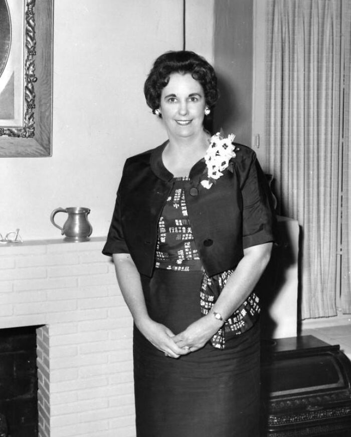 Black and white photo of a LA woman from 1962 dressed elegantly with a floral corsage, smiling indoors by a fireplace.
