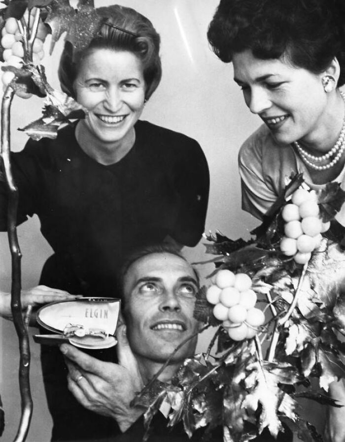 Two LA women in 1962 smiling and interacting with a man holding an Elgin watch amidst decorative grapevine leaves.