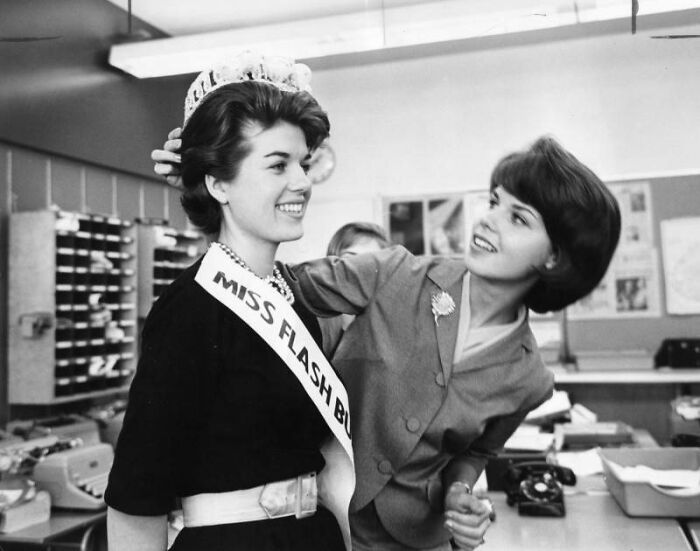 Two LA women in 1962 office setting, one wearing a sash and crown, smiling and enjoying the moment together.