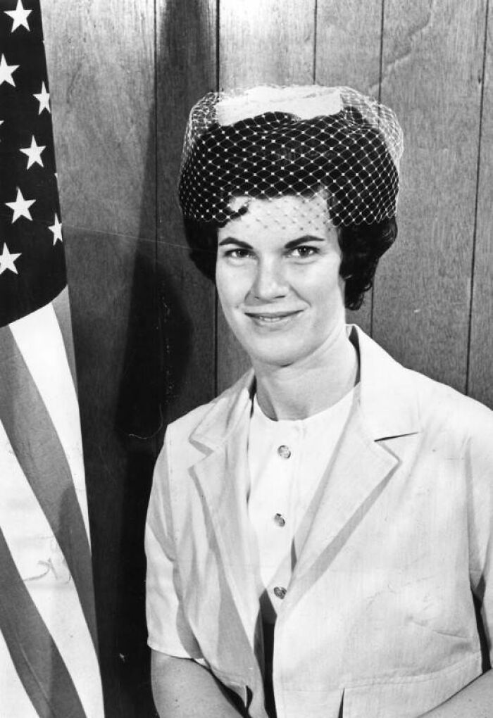 LA woman in 1962 wearing a mid-century outfit and netted hat, posing confidently beside an American flag indoors