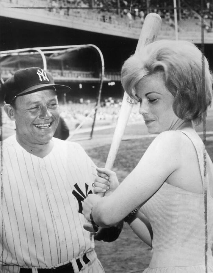 Woman holding a baseball bat receiving batting tips from a New York Yankees player in a vintage stadium setting, LA women 1962.