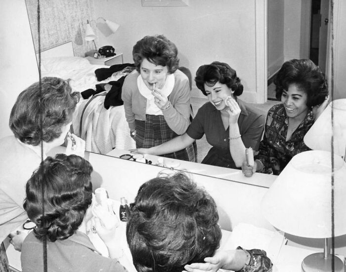 Three LA women laughing and applying makeup together in front of a bedroom mirror, living their best lives in 1962.