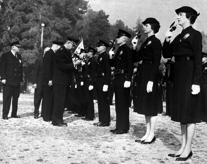 Women in 1962 LA police uniforms standing in formation with pistols during a formal inspection outdoors.