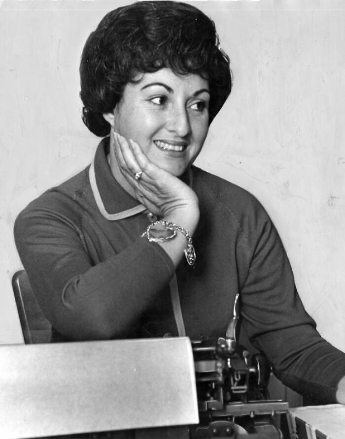 Smiling LA woman from 1962 sitting by a typewriter, wearing a bracelet and looking off to the side thoughtfully.