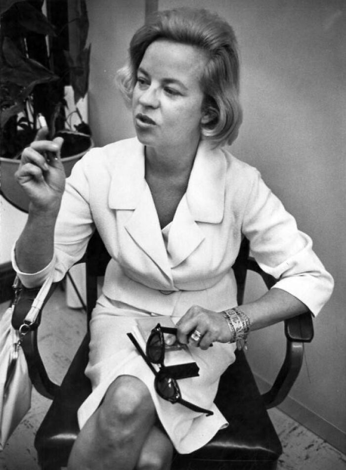 Black and white photo of an LA woman in 1962 wearing a white outfit, holding sunglasses, and sitting in a chair indoors.