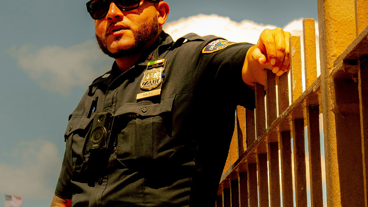 Prison officer in uniform and sunglasses leaning on a gate, reflecting on Epsteinu2019s death with experienced suspicion.