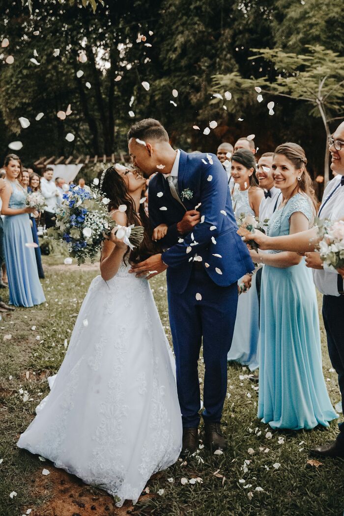 Bride and groom kissing outdoors with wedding party throwing petals, highlighting performative behaviors in society.