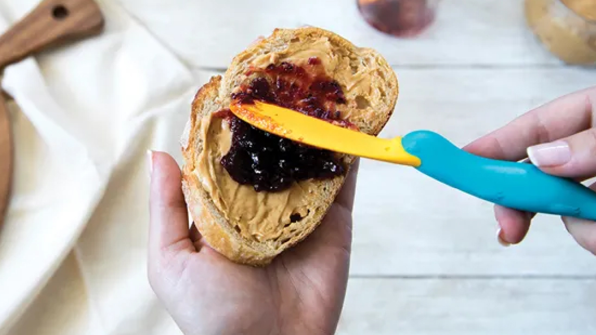 Hand spreading jelly on peanut butter toast with a fun and functional blue and yellow butter knife in kitchen setting