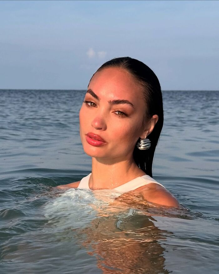 Miss Universe winner posing in the ocean with wet hair and silver earrings under natural sunlight.