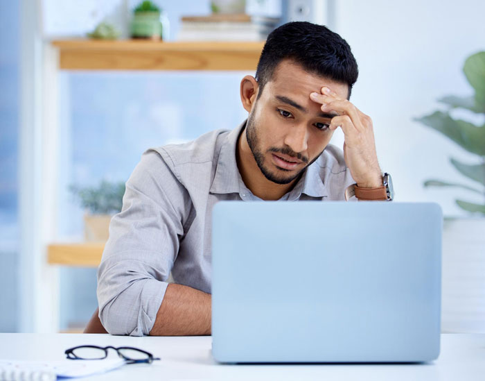 Indian guy looking worried while working on laptop, reflecting concerns about refusing to change name for US client. Indian guy looking worried while working on laptop, reflecting concerns about refusing to change name for US client.