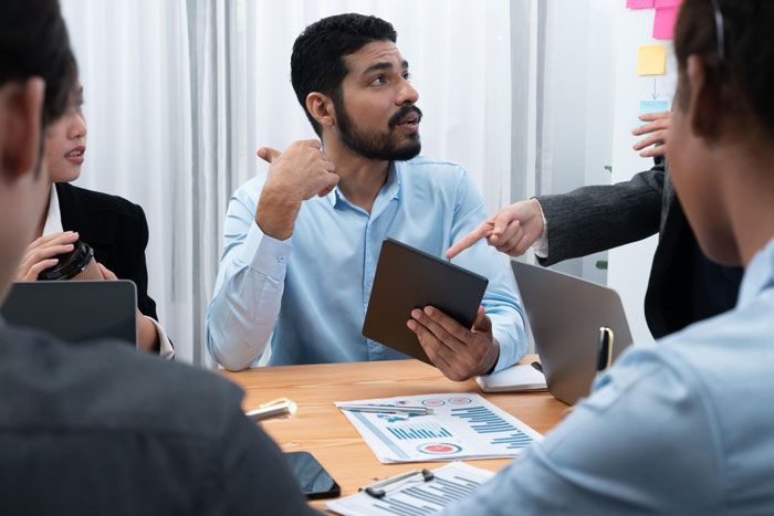 Indian guy in a business meeting holding a tablet, appearing to refuse a request while colleagues discuss career risks.
