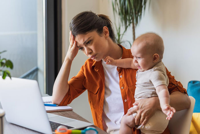 Mother caring for 10-month-old looks stressed while holding baby and working on a laptop in a home setting.