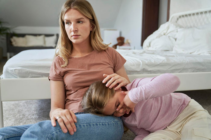 Two teenage girls sitting on the bedroom floor, one comforting the other, highlighting twins overwhelmed by babysitting duties.