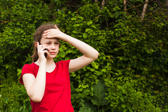 Young girl in red shirt talking on phone outdoors, showing concern in a relationship with siblings and mom context.
