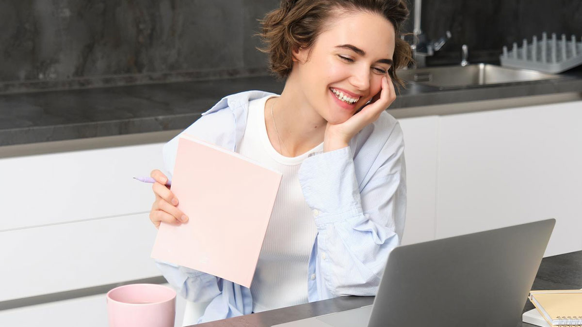Smiling woman working on laptop in kitchen, holding notebook, illustrating woman quits paying rent and apartment dispute.