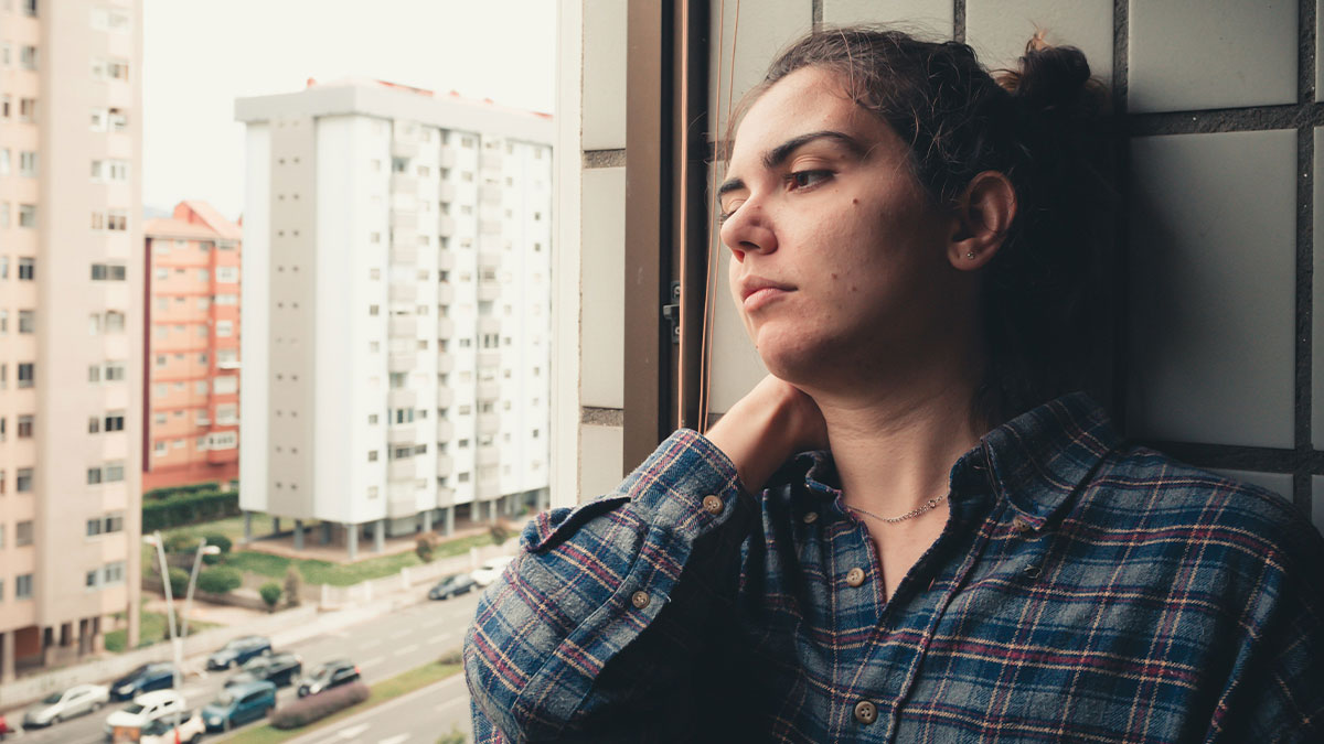Young woman in plaid shirt looking out window at cityscape, capturing the feeling of boring dystopia in modern life.