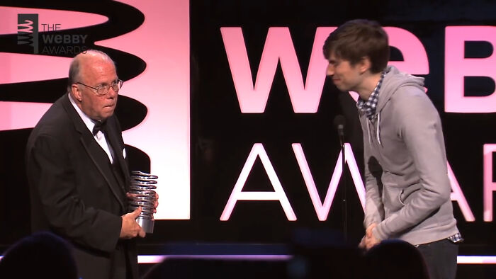 Two men on stage at an awards show, with one holding a trophy and the other speaking at the microphone, capturing savage comebacks.